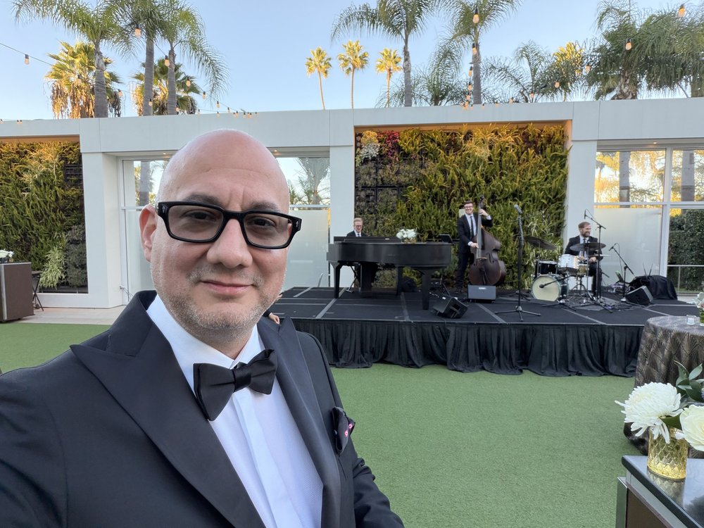 Luis Giraldo in a tuxedo at the Beverly Hilton, with the jazz trio performing on stage behind him in a palm-lined garden