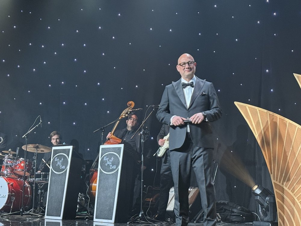 Luis Giraldo on stage at the Beverly Hilton in a black tuxedo, with art deco backdrop and jazz trio behind him